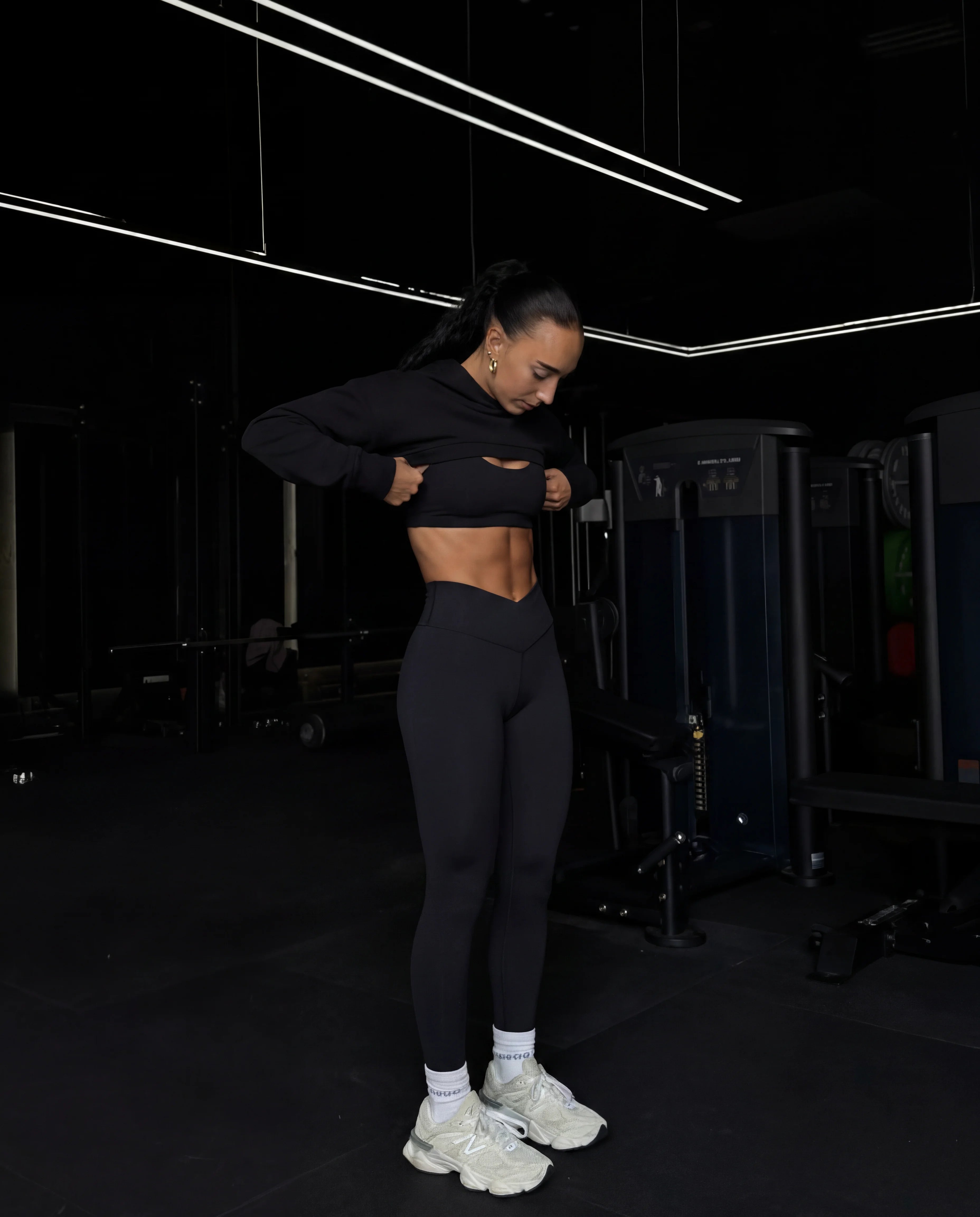Black cropped hoodie and leggings adjusting fit in dark gym