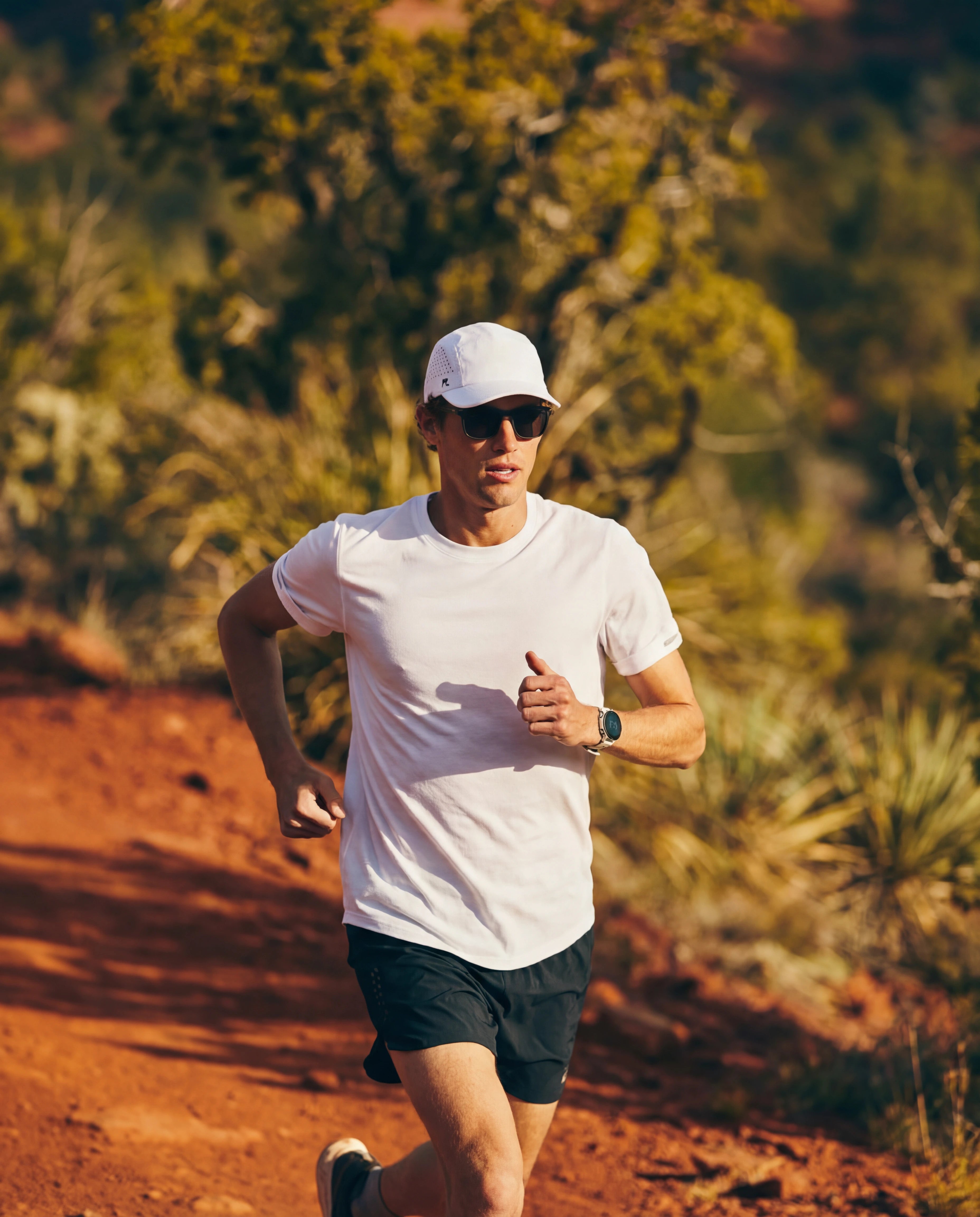White running hat and running sunglasses on man trail running in white tee and navy shorts from Runners Athletics