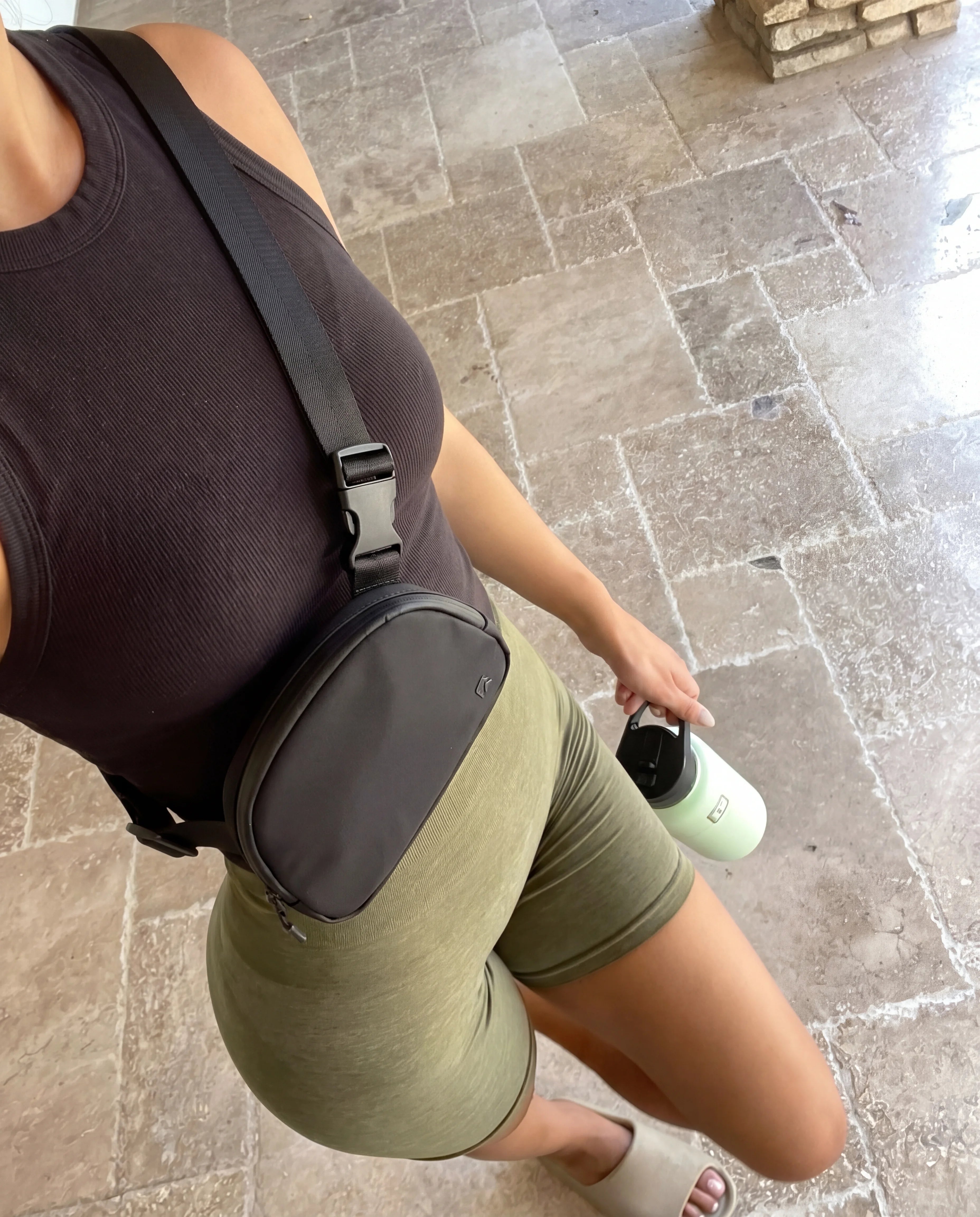Black crossbody sling bag worn over dark tank top and olive biker shorts