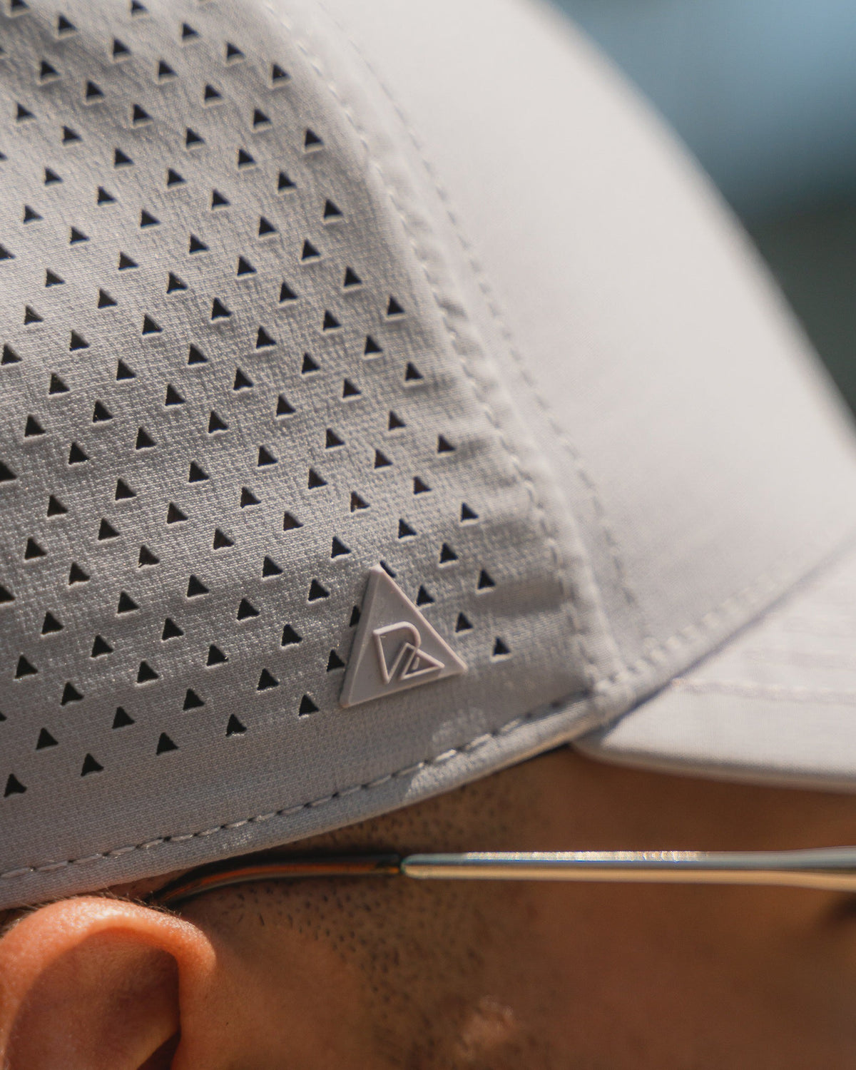 Close-up of a gray athletic hat with triangular perforations and a triangular logo, worn by a person with glasses.