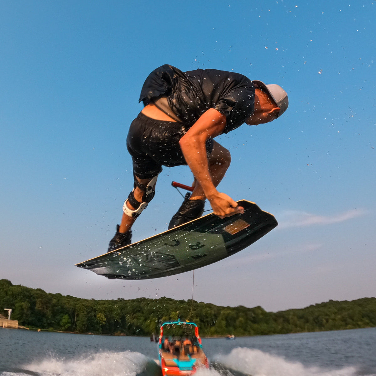 Wakeboarder in mid-air, grasping the board, with water droplets, over a lake with a boat and trees.